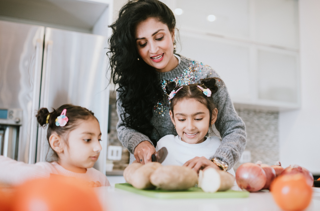 a mother carefully helps her 2 school aged daughters cut potatoes while having fun preparing food in the kitchen