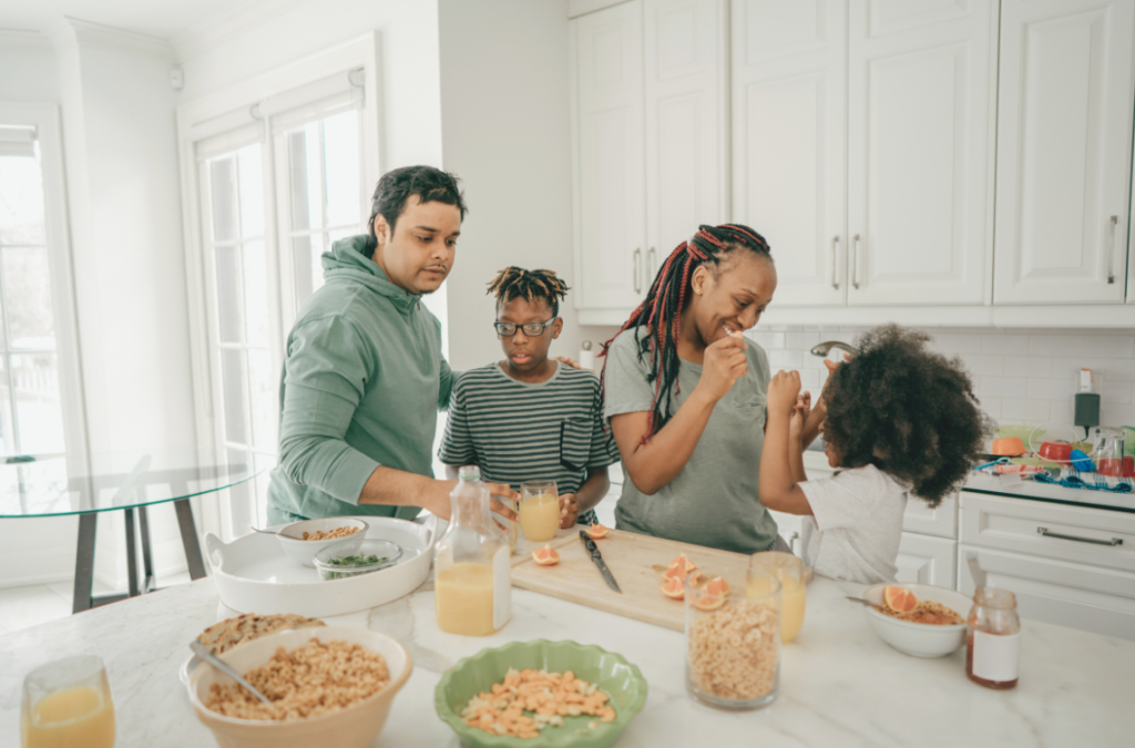 Multiracial family (mom, dad, son, & daughter) preparing breakfast with fruit, orange juice, and cereal at the kitchen counter