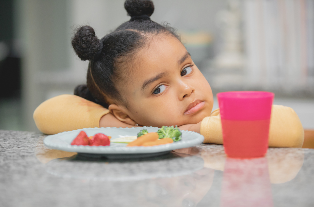 child with head on arms on the table looking disappointed staring at a vegetable plate